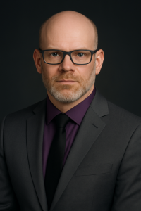 Photo of James Kilmer-Lawler wearing a dark grey suit and tie with a burgundy shirt